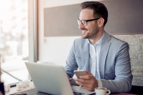 Un manager spécialisé sur la RSE souriant habillé en chemise est assis à une table avec un téléphone à la main et son ordinateur et café posés sur la table